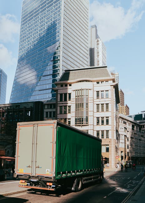 A large green-moving truck with a covered cargo area is parked on a city street in front of modern glass and stone office buildings, which are reflected on the curved glass facade of one tall skyscrap