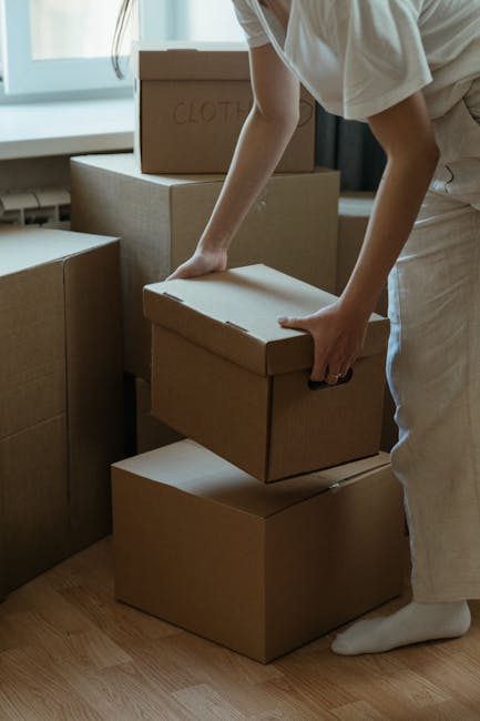 A person dressed in light-coloured clothing is seen lifting a medium-sized cardboard box, marked with the handwritten word 'CLOTHES' on the top, which is part of a larger stack of similar boxes. The b