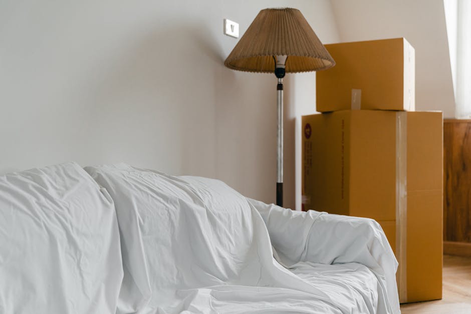 A person dressed in light-coloured clothing is seen lifting a medium-sized cardboard box, marked with the handwritten word 'CLOTHES' on the top, which is part of a larger stack of similar boxes. The b