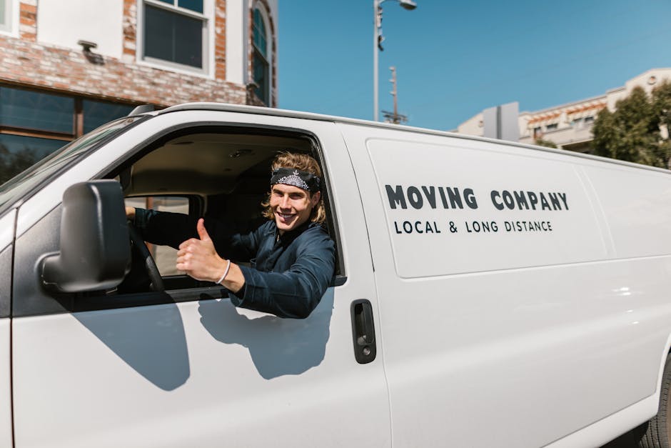A young male mover with long, wavy hair wearing a black headband and dark clothing is sitting in the driver's seat of a white removal van labeled 'MOVING COMPANY, LOCAL & LONG DISTANCE,' parked outdoo